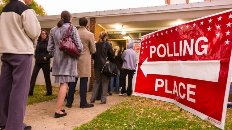 A red sign in the foreground points a line of voters toward a polling place.